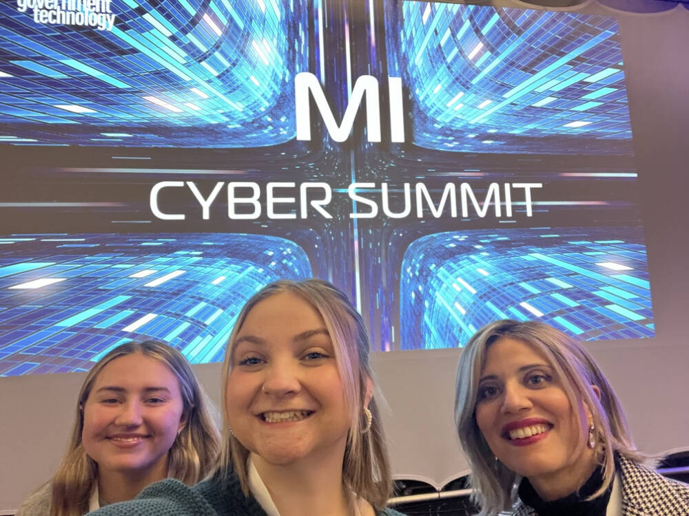 Cybersecurity students, Audry Douglas and Sophi Ernst, along with Dr. Samah Mansour smile for a selfie at the MI Cyber Summit, with a digital-themed event backdrop displaying "MI Cyber Summit" in bold text.